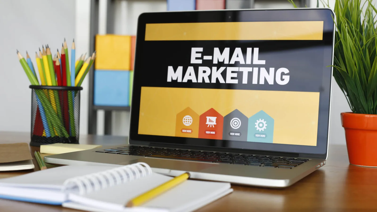 A laptop on a desk with an email marketing screen, beside coloured pencils, a notebook and a plant